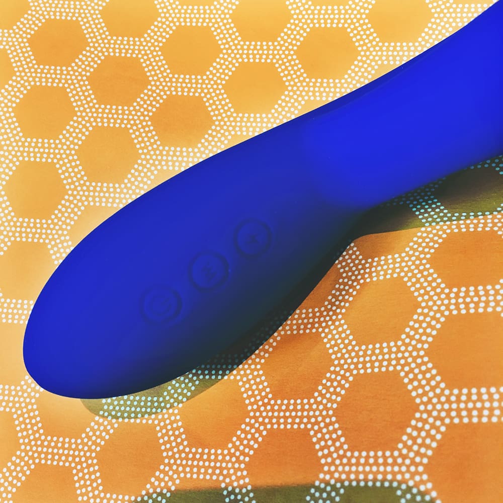Photo of blue rabbit-style vibrator on yellow heagonal pattern background. Photo shows operational buttons - e-stim, vibrate, and power
