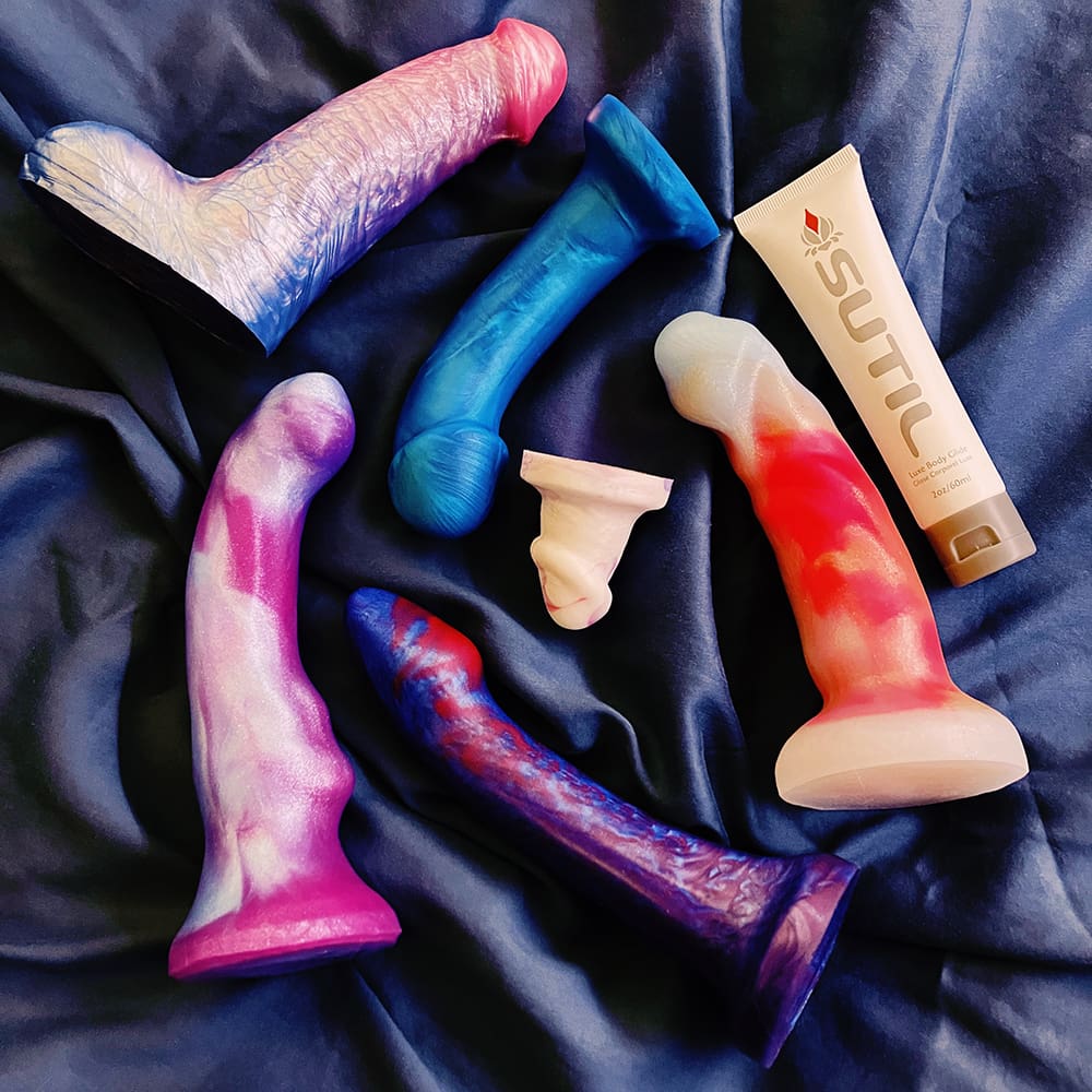 photograph of an array of 6 dildos in mostly white, purple, blue, and pink collocations on a blue cloth background, one bottle of Sutil lube